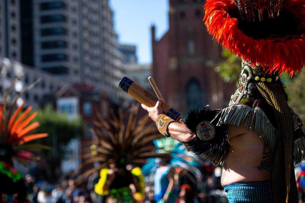 A close-up of hands holding a tubular wooden instrument and a drumstick, with an elaborate headdress in the background.
