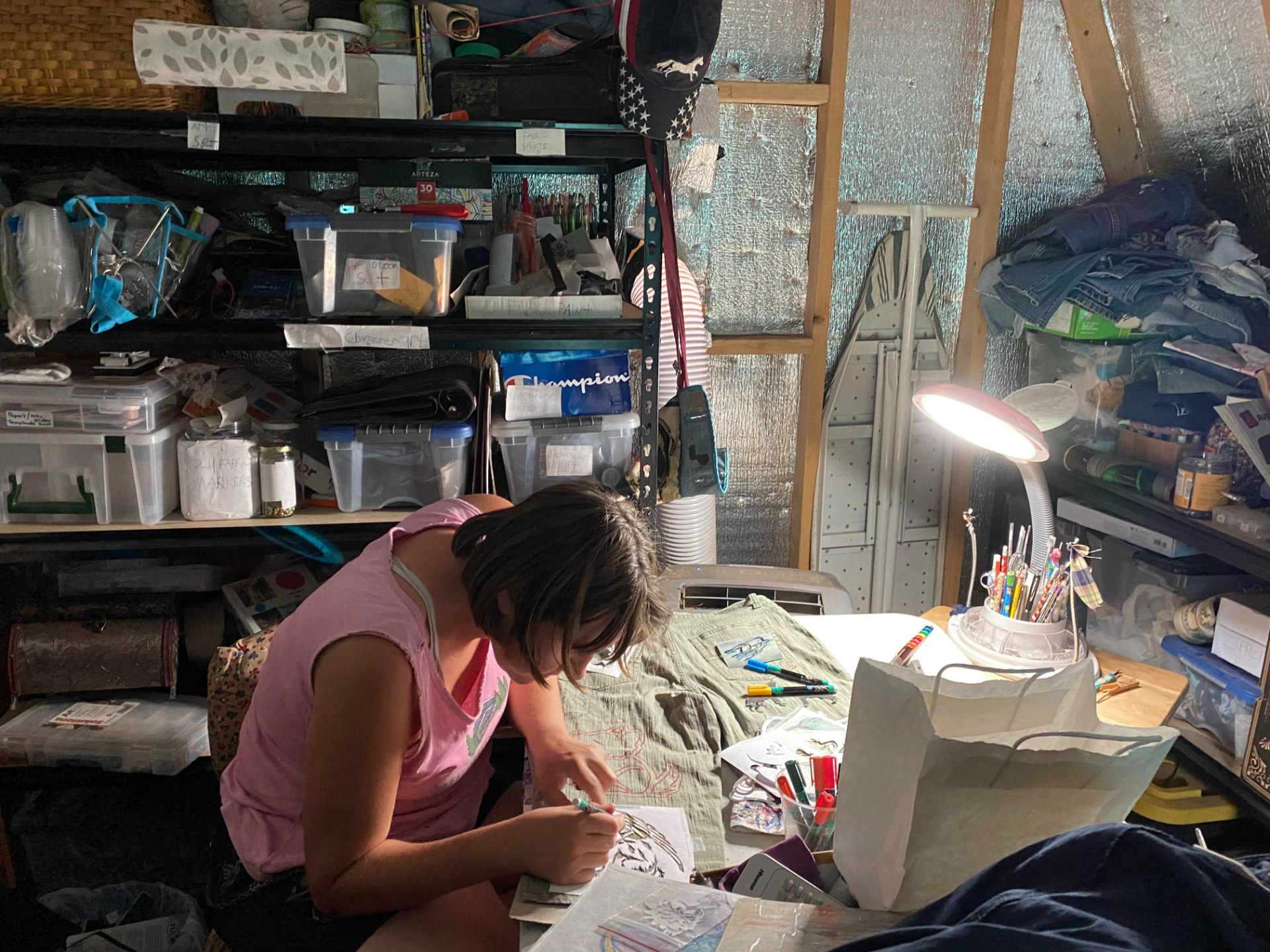 A woman bends over some crafting work in a shed, with a lamp hovering over her work.