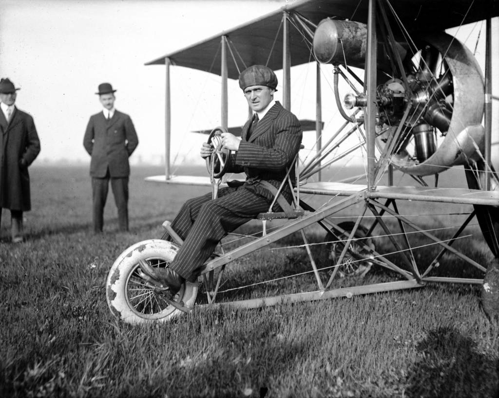 Black and white photo of a man sitting on an early model airplane.