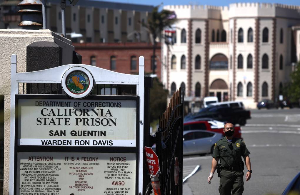 A man in uniform stands next to a sign that says "California State Prison, San Quentin," with the castle-like facade of the prison behind him.