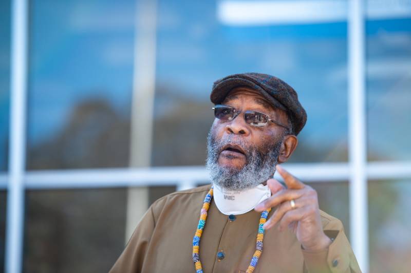Amos Brown, in a hat, beard, glasses, and green, blue and yellow necklace, raises his hand mid-speech, the windows of a building behind him.