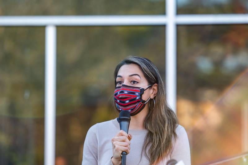 A woman with long brown hair and a red and black woven masks holds a microphone in front of windows reflecting trees.