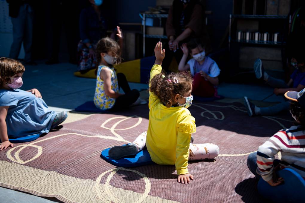 A little kid in a bright yellow sweater, sitting in the sun on a mat amid other little kids with their hands up, raises her hand.