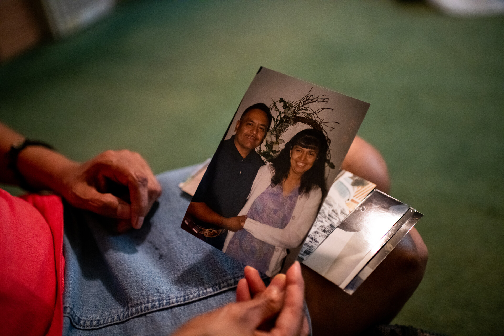 A hand in a lap holds up a photo of a smiling couple, holding hands, in casual dress in front of a round wall hanging of twigs and blooms.