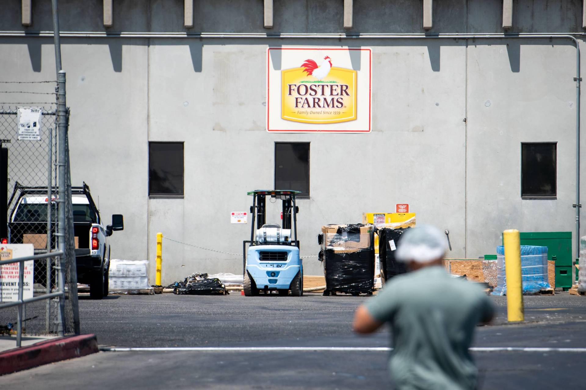 A man in a plastic hairnet and a T-shirt walks toward a concrete building and a forklift beneath a Foster Farms logo.