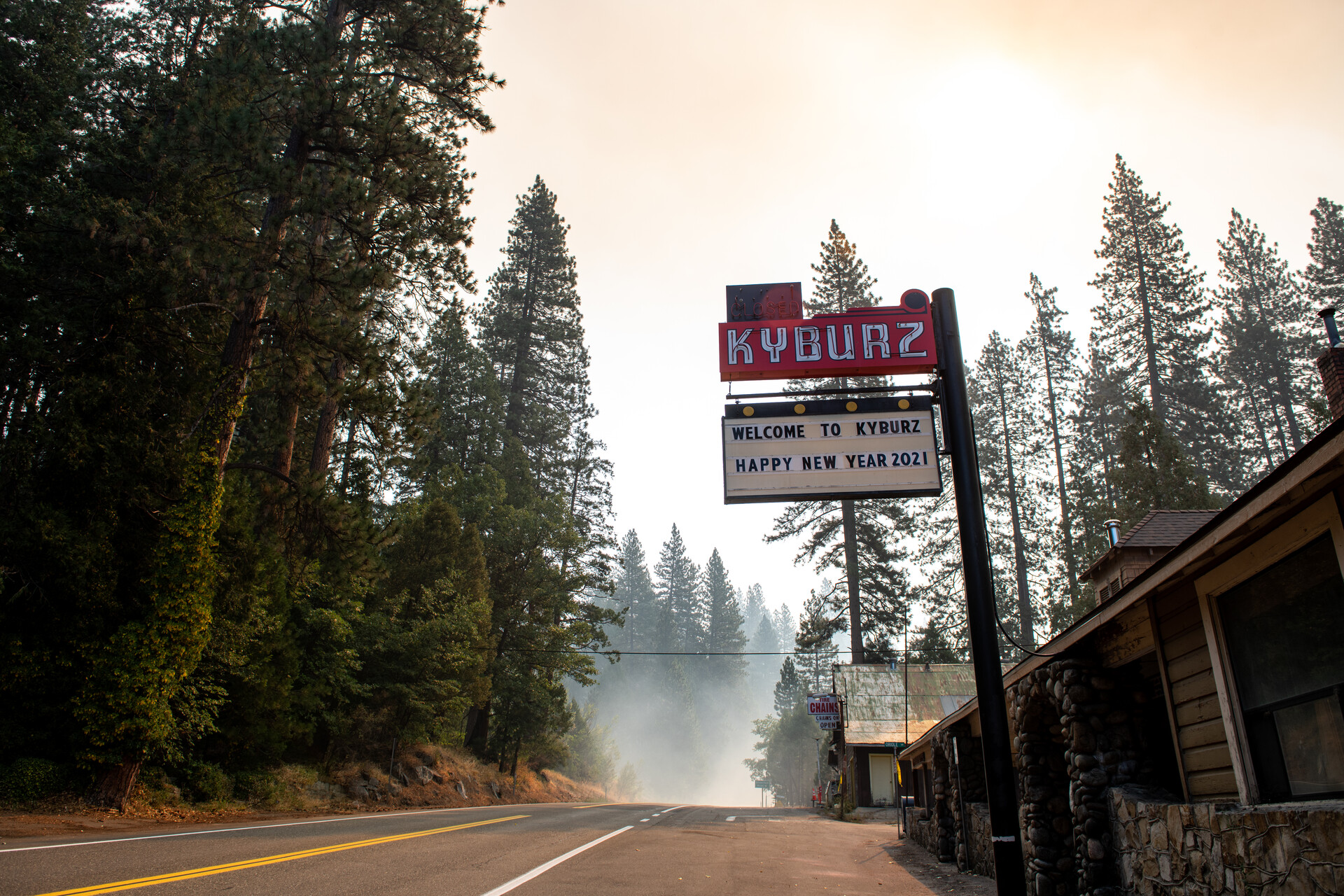 A red and white sign on a tall pole along a two-lane road, surrounded by tall fir trees, with smoke haze in the distance.