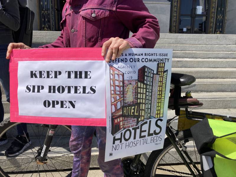 A person holds two signs in front of a set of outdoors steps. One says, "Keep the SF Hotels Open," and the other shows a drawing of high-rises and the word "Hotels."