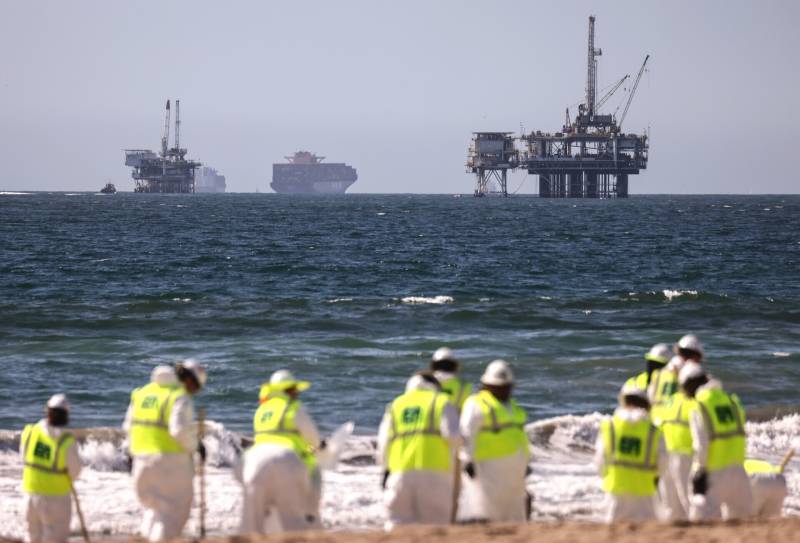 A group of white-suited people in white hard hats and neon yellow jackets work on the beach, with oil platforms in the distance in the ocean.