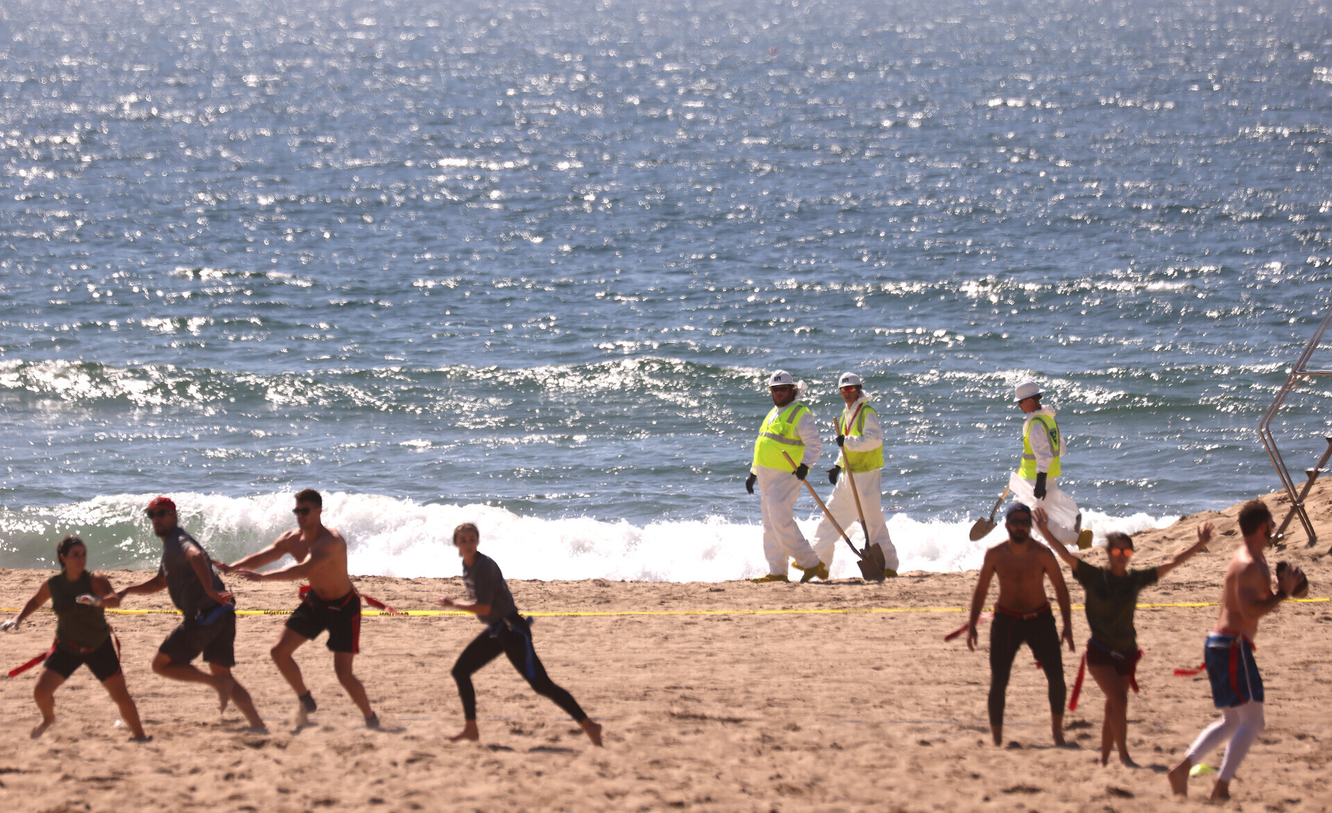On a sunny beach along the sunlit ocean, three people dressed all in white with neon yellow jackets and white hard hats carry tools beyond a group of people wearing shorts, one holding a football.