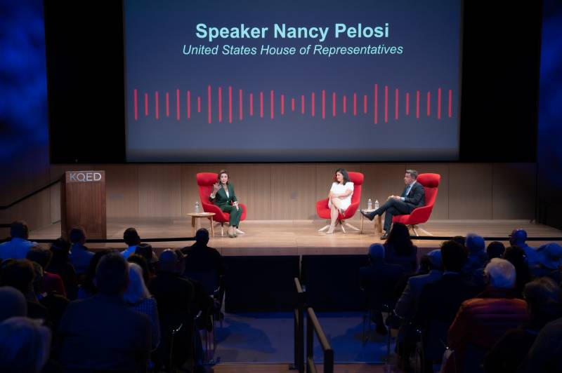 A stage in a darkened auditorium, with three red chairs for, L-R, Nancy Pelosi, Marisa Lagos, and Scott Shafer. A large screen in the background says "Speaker Nancy Pelosi, United States House of Representatives."