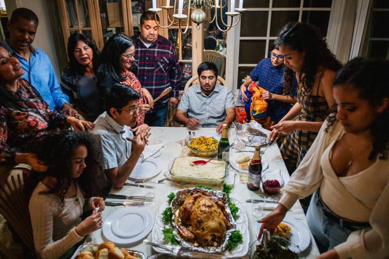 Family members stand around a table with many Thanksgiving dishes.