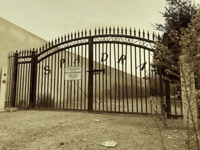 A wide and ornate driveway get to Spadra cemetery which has letters spelling S P A D R A across the bars. The photo is tinted sepia and yellow.