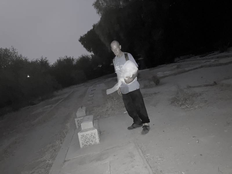 A bald man in a baggy t-shirt and loose pants standing at the edge of a sidewalk in the center of the frame with trees in the background. The photo appears to be taken in a "night" mode, and his eyes are shining unnaturally.