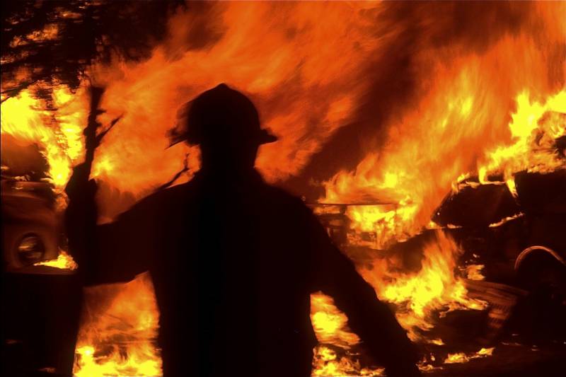 A person in a hard hat moves in silhouette of an entirely orange-and-black scene -- nothing is discernible other than flames, and maybe the outline of a car wheel.