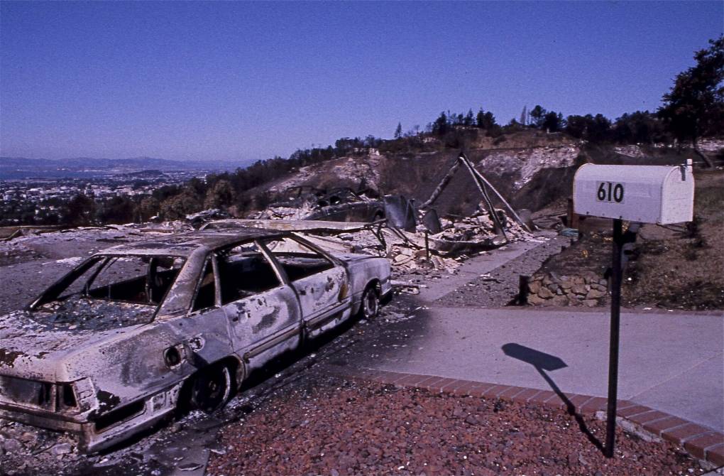 Grainy image shows a pristine mailbox with the address 610 overlooking a singed driveway that leads to a charred car with wildfire rubble and the ridge of the East Bay Hills in the distance.