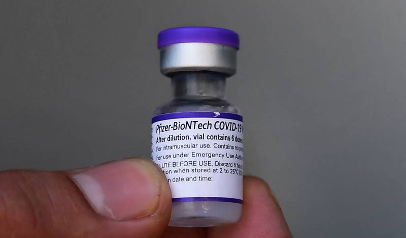 A finger and thumb hold up a purple-capped Pfizer vaccine vial.