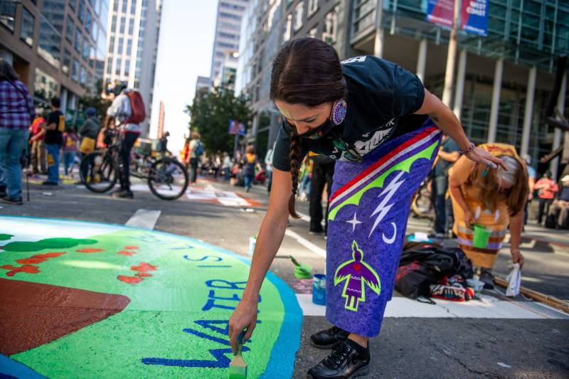 A young woman in a purple skirt paints with green on a San Francisco street as part of a climate rally