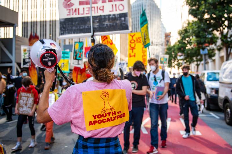 A woman stands back to camera with a "youth vs apocalypse" sign on her back and holding a bullhorn.