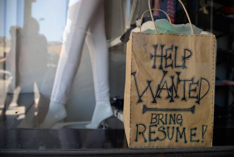 A shopping bag with the message "Help Wanted. Bring Resume!" next to the legs of a mannequin is displayed in a shop window.