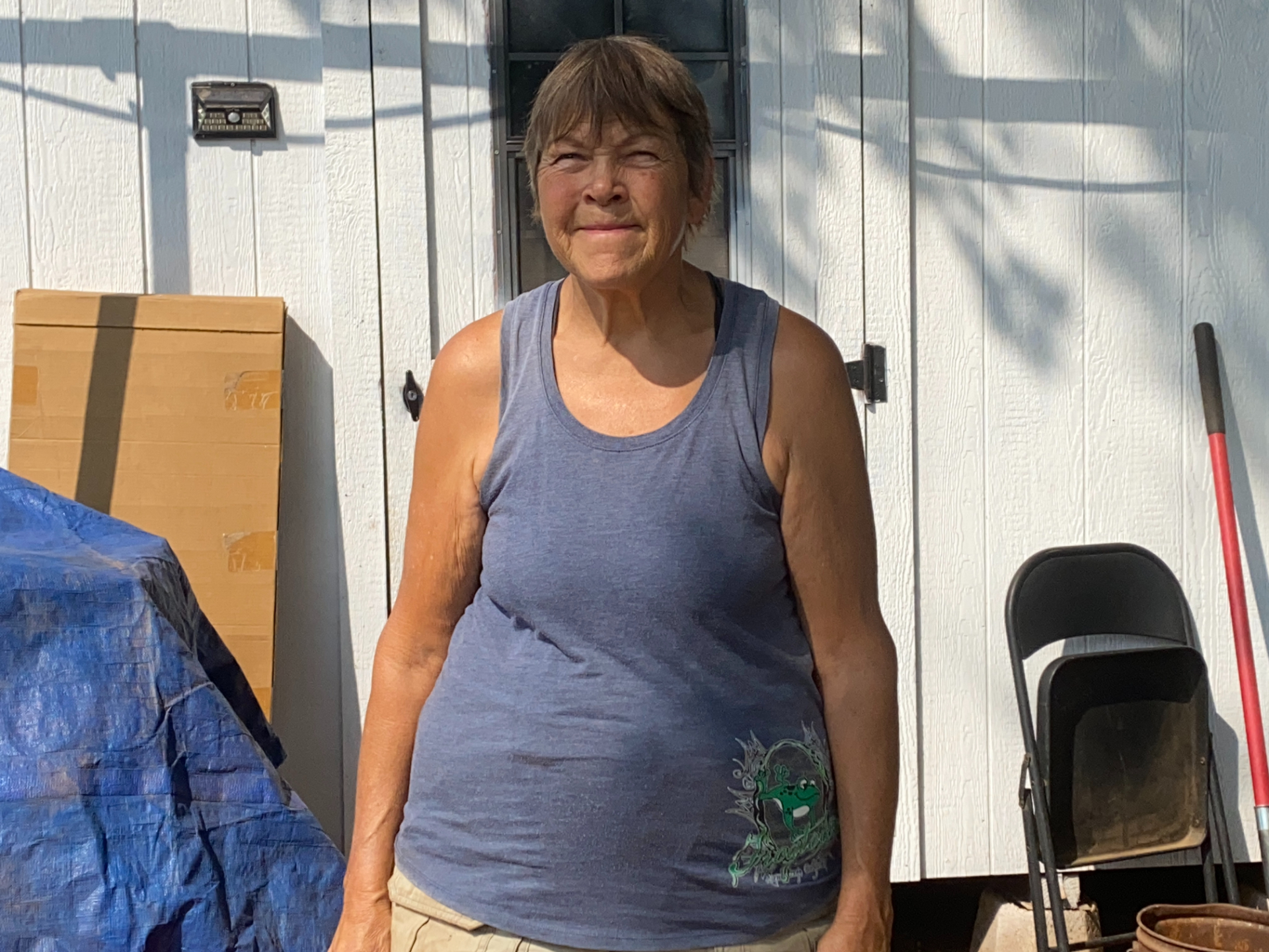A woman in a tank top stands in front of a storage shed, with a slight smile and eyes closed in the bright sun.