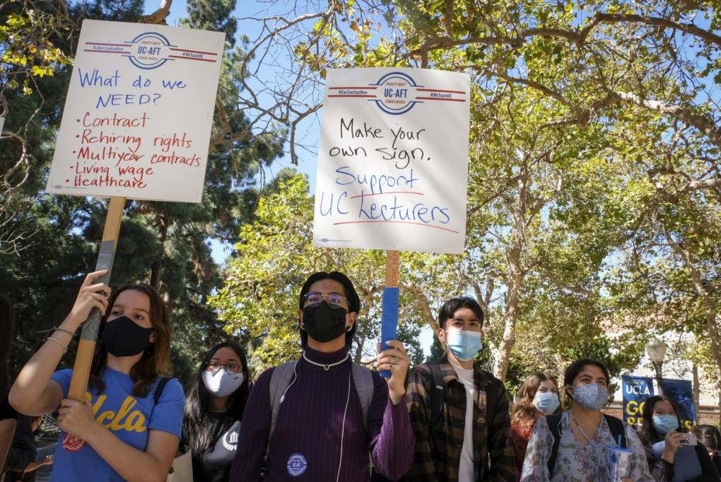 Masked students walking beneath leafy trees hold signs. One says, "Support UC Lecturers." Another says, "What do we need? Contract, rehiring rights, multiyear contracts, living wage, healthcare."