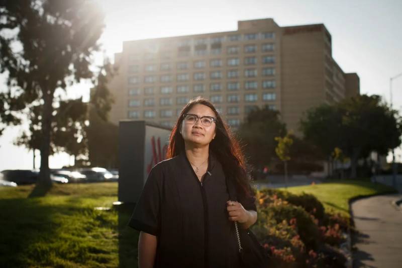 A woman with long hair and glasses, lightly smiling and looking above the camera, stands in front of a large hotel, with sun cresting its corner behind her, looking calm and powerful.