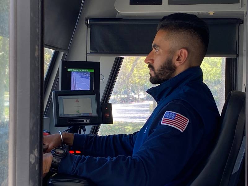 A man wearing a blue work jacket with an American flag patch on the left shoulder sits at the controls of a train, looking through the windshield.