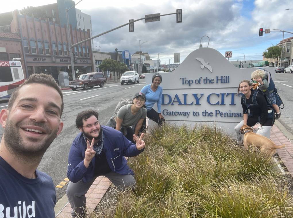 Five people look joyous and somewhat tired while standing next to a sign that says "Daly City: Gateway to the Peninsula"