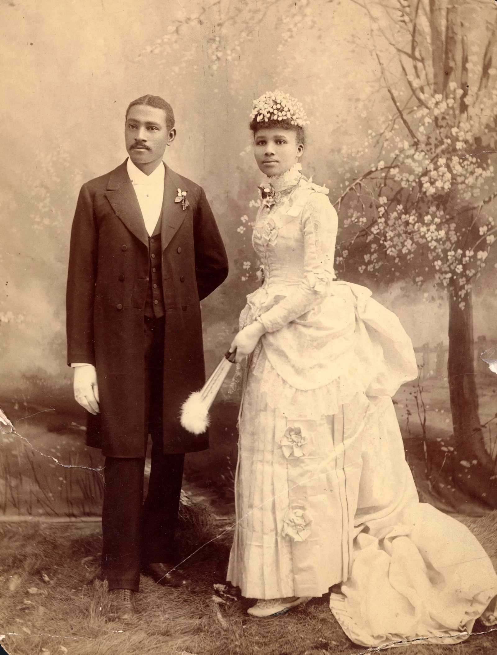 Sepia-tone portrait of an elaborately dressed couple, one in a three-piece suit and the other in a white dress with a bustle, holding a fan.