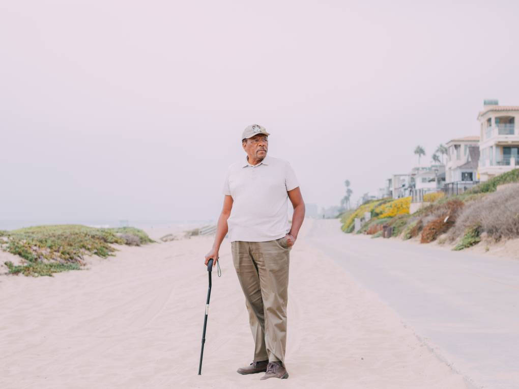 A man in a ball cap and a polo shirt poses on the sand, one hand resting on the head of a cane.
