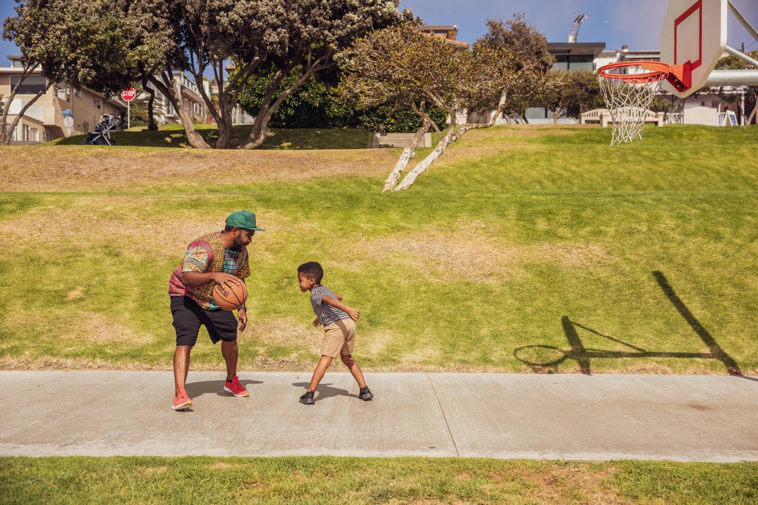 A man dribbles a basketball on a sidewalk between two green lawns with his son, who looks about 5.