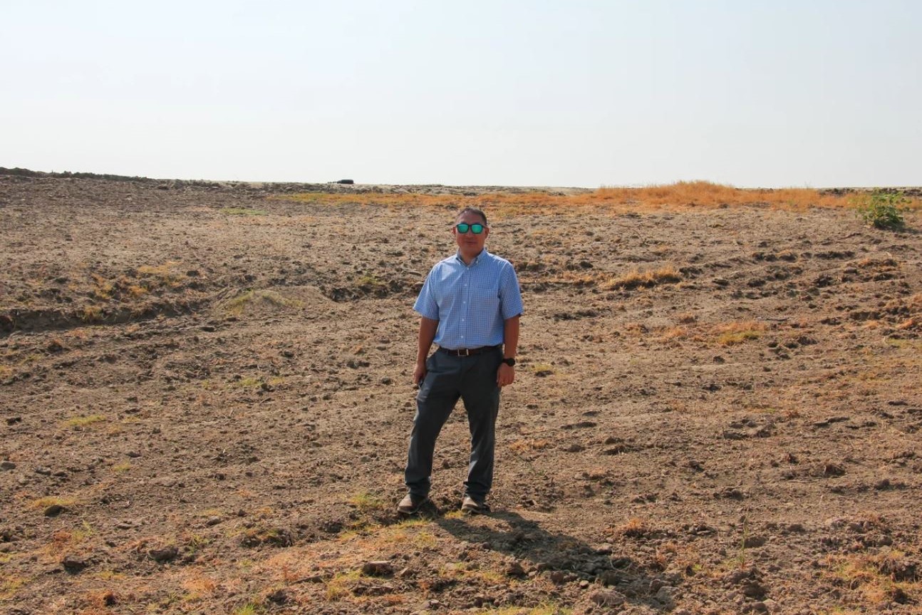 Aaron Fukuda stands in a dry and empty field, wearing reflective sunglasses and looking at the camera.