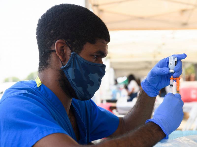 A man with a face mask, purple gloves and blue short-sleeved scrubs fills a hypodermic needle from a vial.