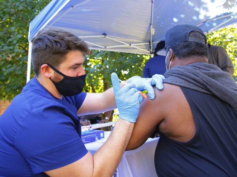 A man in blue scrubs, a mask, and blue gloves gives a man in a black T-shirt and baseball cap a shot in his upper arm beneath a tent outdoors amid trees.