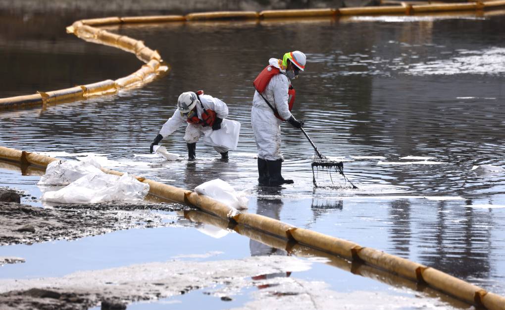 Two people in white suits, orange life jackets, boots and gloves rake oil amid yellow inflatable booms in ankle-deep water.