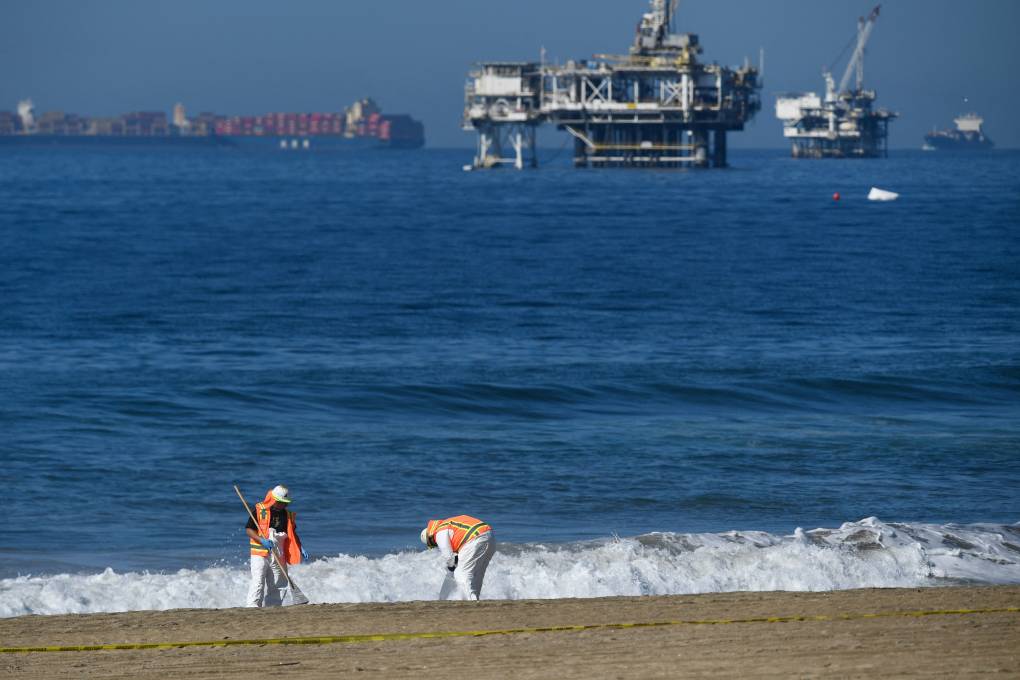 Two people in white hazmat suits and orange life vests pick up oil on a sandy shore, with an oil platform (?) and a cargo ship visible in the distance.