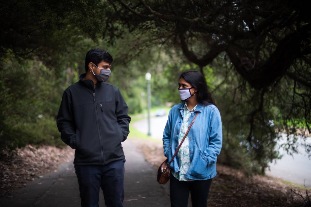 A man and woman walk through a park wearing COVID masks. He's in a black jacket and pants, she's in a blue jacket and white top.