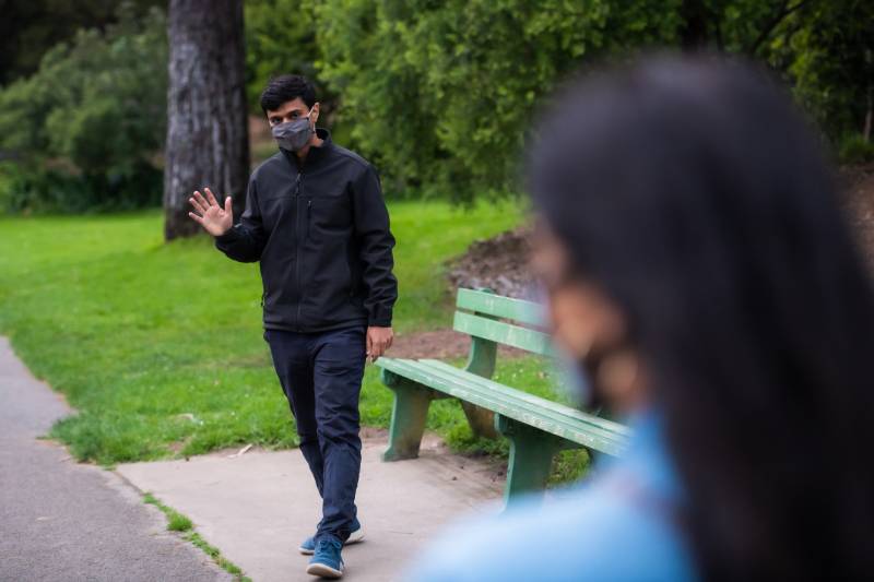 A man in a black jacket and dark pants waves as he walks. He has black hair and wears a COVID mask, with grass and trees behind him. A woman is in the front right of the frame turned away from the camera, blurred.