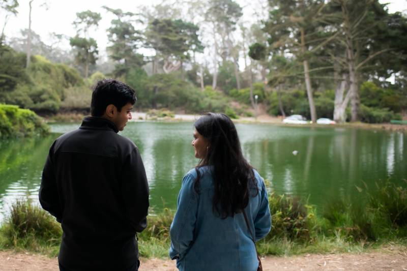 A man and woman stand, backs turned to the camera, in front of a pond. The couple are turned to each other as if they're talking.
