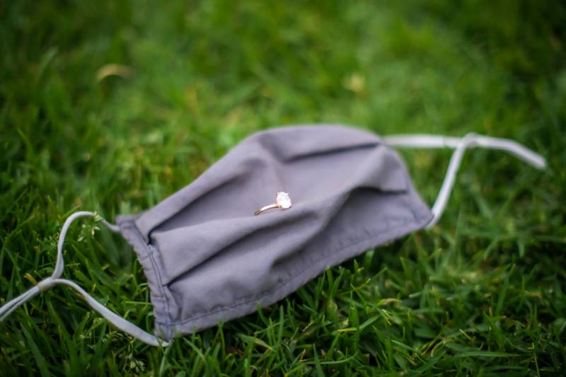 A portrait of an engagement ring sitting atop a black covid mask, laying on grass. 