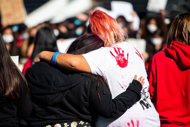 Seen from behind, two students hug one another. On the right, a student in a white tshirt with red and blonde hair, the tshirt has red blood-colored palm prints. The student on the left is in a black hoodie. They are standing in a crowd.