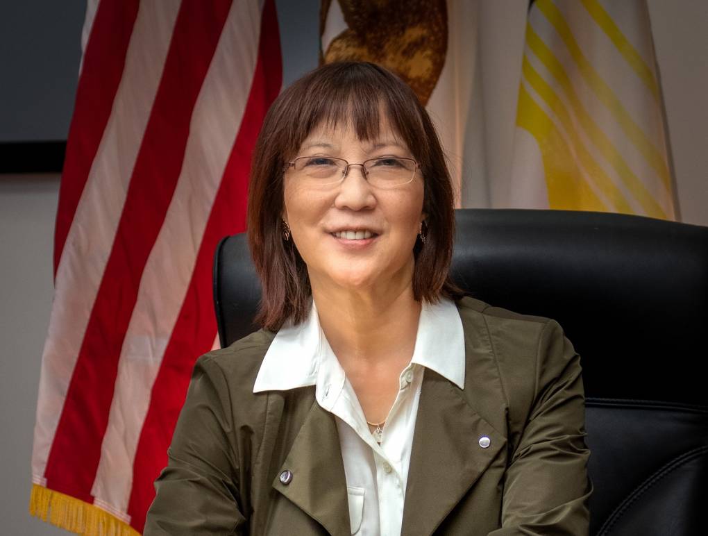 A woman, smiling and wearing glasses, an olive blazer, and a white blouse with a wide collar sits, at a desk in front of an American flag.