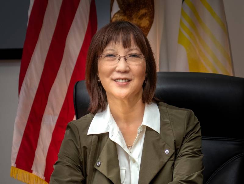 A woman, smiling and wearing glasses, an olive blazer, and a white blouse with a wide collar sits, at a desk in front of an American flag.