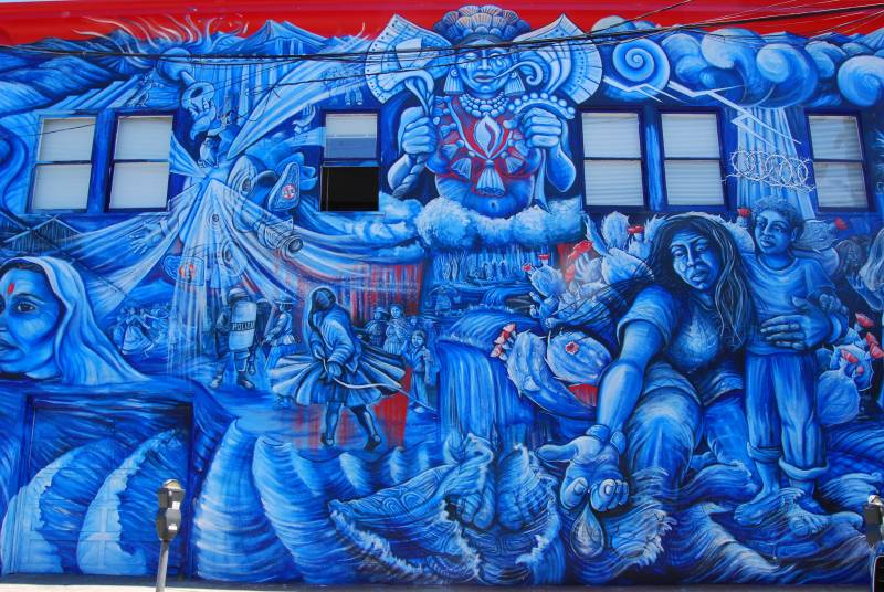 An intricate wall mural painted in broad strokes of blue. At the center is Chalchiuhtlicue, the Aztec goddess of lakes and streams. In the background, there are women from Bolivia, women from India, women from the Mexico-U.S. border — all standing together. The figure of La Llorona herself is in the foreground, holding a child, and with a single tear on her cheek. Her hand is outstretched toward the viewer.