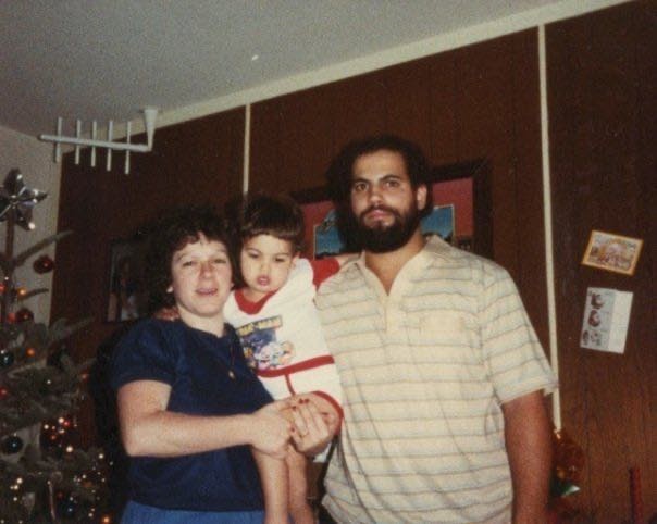 A photo with an older, vintage camera look, inside a room with a Christmas tree on the left, white ceiling and wood paneling behind a family of three. The mother is White with brown straight hair, holding her son who is in what look like white and red trim pajamas with a Pac Man logo. The father on the right is Puerto Rican, sporting a bit of a serious face, with a dark brown beard and what the child in the middle, now grown up, described as an Afro. The father is in a striped beige polo, and the boy, who is looking down, has his arm around his neck 