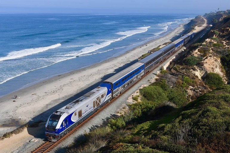 An aerial shot of a long, silver-and-blue train on tracks, with only a few yards of beach between it and ocean waves, with low, green hills on the immediate opposite side.