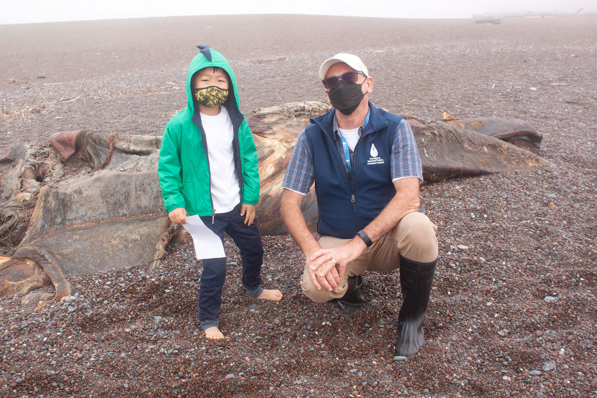 Five year old child in green jacket poses on a pebbly beach with older man in a blue vest, white baseball cap, and knee high rubber boots. Behind them is the decomposing carcass of a whale.