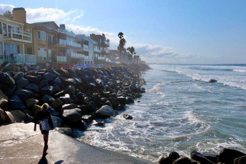 Large rocks separate a row of beachfront homes from the rising tide. A surfer looks on from a pier in the foreground.