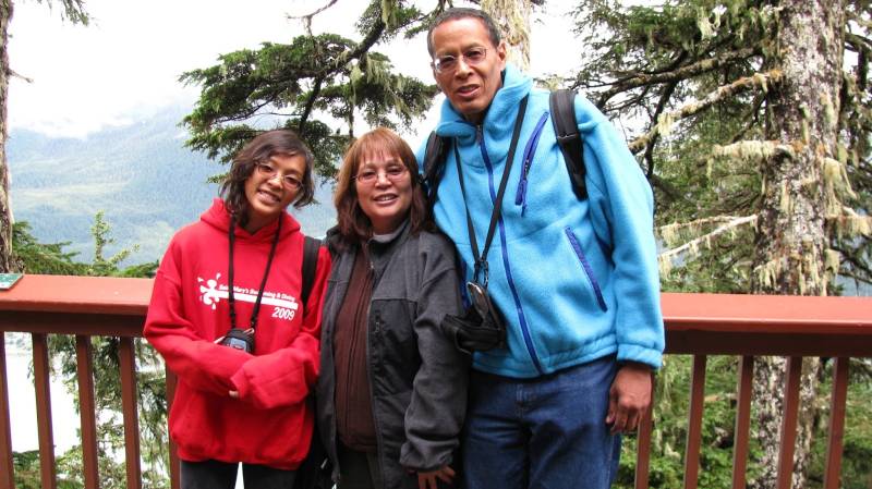 A multiracial family stands together outside, with a copper-colored guardrail, trees and a forest in the background. The father is tall in a light blue hoodie, with an arm around the mother who is in a black jacket and smiling, with their daughter on the far left in a red hoodie and glasses, also smiling. They appear to be on a tourist trip. 