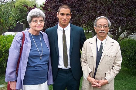 A family of three stands well-dressed in front of a few trees, outside on the grass. On the left, the mother is Jewish in a violet jacket and short gray hair, to the right is the father who is Japanese, sporting a brush-like mustache and in a suit and trenchcoat, with a slight smile that may be characterized as a smirk, and their son is in the center, well dressed in a suit with a close cropped haircut of his black hair, himself smirking.